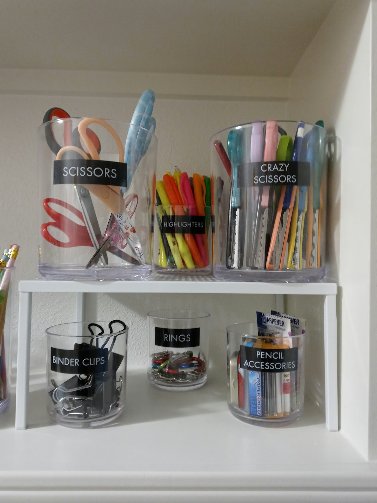 Clear plastic containers labeled with office supplies on a white shelf.